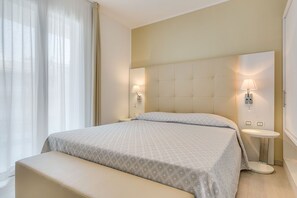 Une chambre d’hôtel avec un grand lit, des tables de chevet, un banc et une tête de lit agrémentée de lampes fixées au mur.