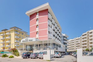 Un hôtel à plusieurs étages, doté d’une façade rouge et blanche bien visible, d’une entrée couverte et d’un parking où sont stationnées plusieurs voitures.