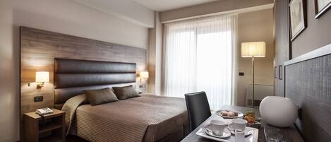 A hotel room with a large bed, bedside lamps, a desk with a tray of breakfast items, and a large window with curtains.
