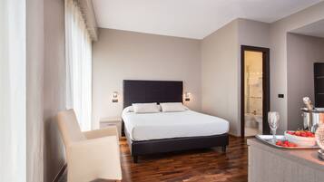 Une chambre moderne avec un grand lit, une table de chevet, une chaise et un petit coin cuisine équipé d’un panier de fruits et d’un verre à vin.