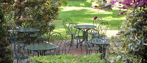 Un patio jardín con mesas y sillas de metal, rodeado de abundante vegetación y un banco al fondo.