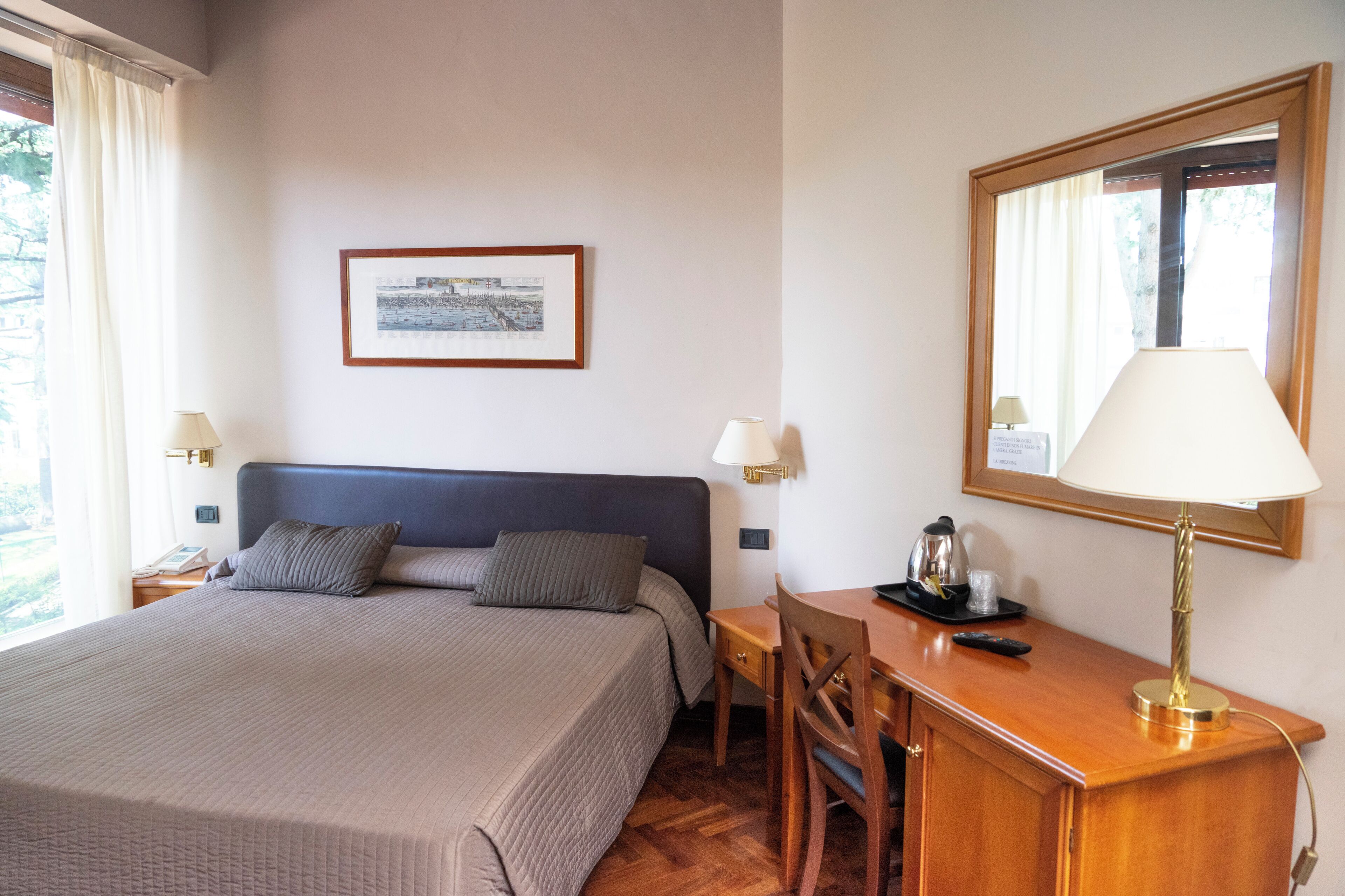 Ein Hotelzimmer mit Bett, Nachttischen, einem Schreibtisch, einem Sessel, einer Lampe, einem gerahmten Bild an der Wand und einem Fenster mit Vorhängen.