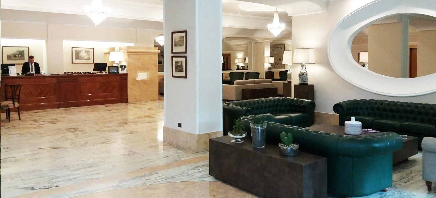 A hotel lobby with a reception desk, green upholstered sofas, a marble floor, and a large mirror.
