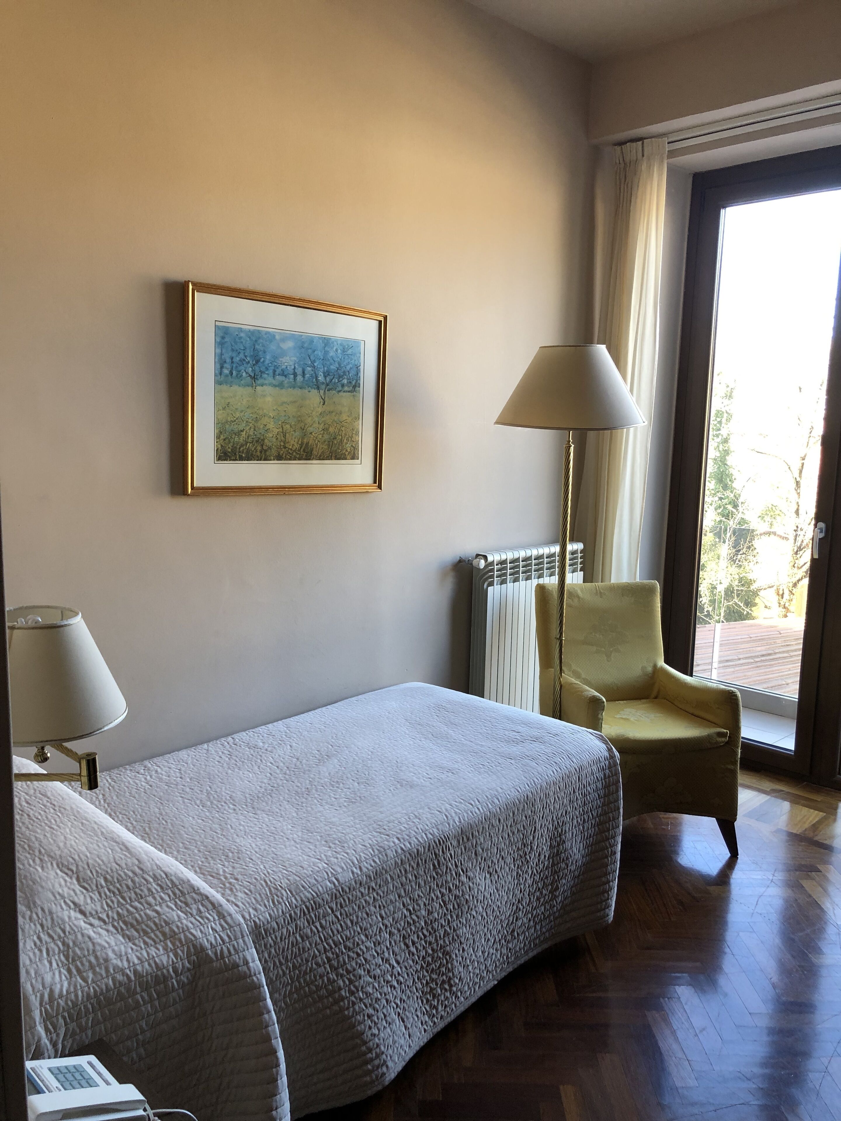 Una habitación de hotel con una cama, una silla, una lámpara, un radiador y una ventana con cortinas.