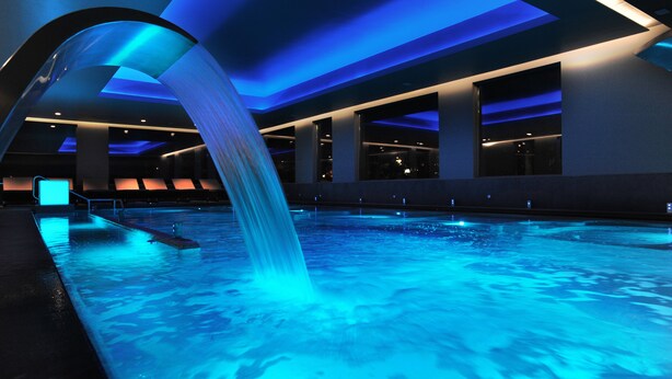 A modern indoor pool with a blue-lit waterfall feature, surrounded by a spacious, well-lit area with seating. - Hotel San Giorgio  Heaven Spa (Fiuggi)
