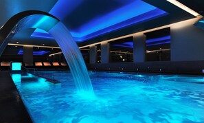 A modern indoor pool with a blue-lit waterfall feature, surrounded by a spacious, well-lit area with seating.