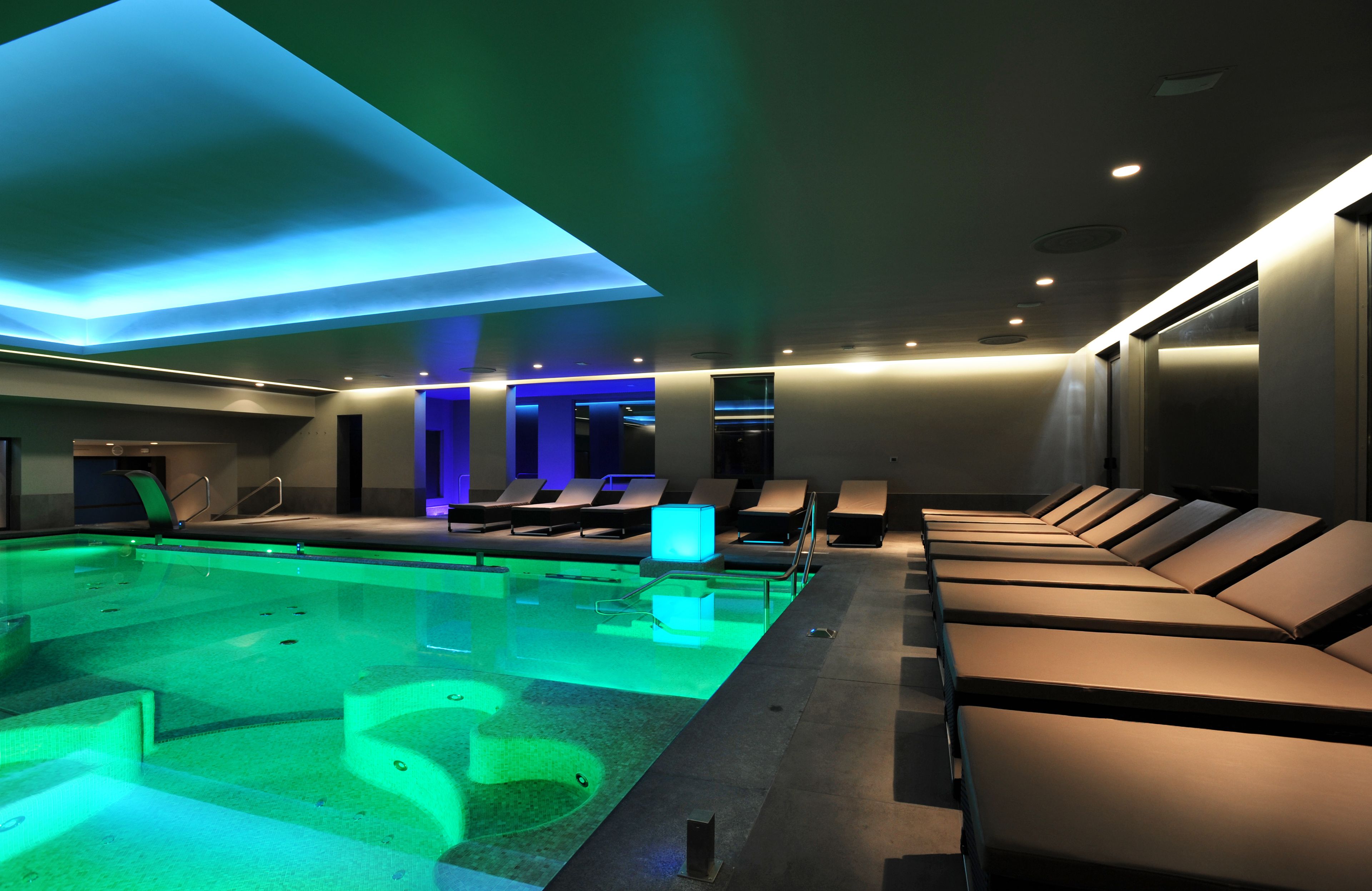Un área de piscina interior moderna con una piscina larga y rectangular, sillones de descanso e iluminación azul en el techo.