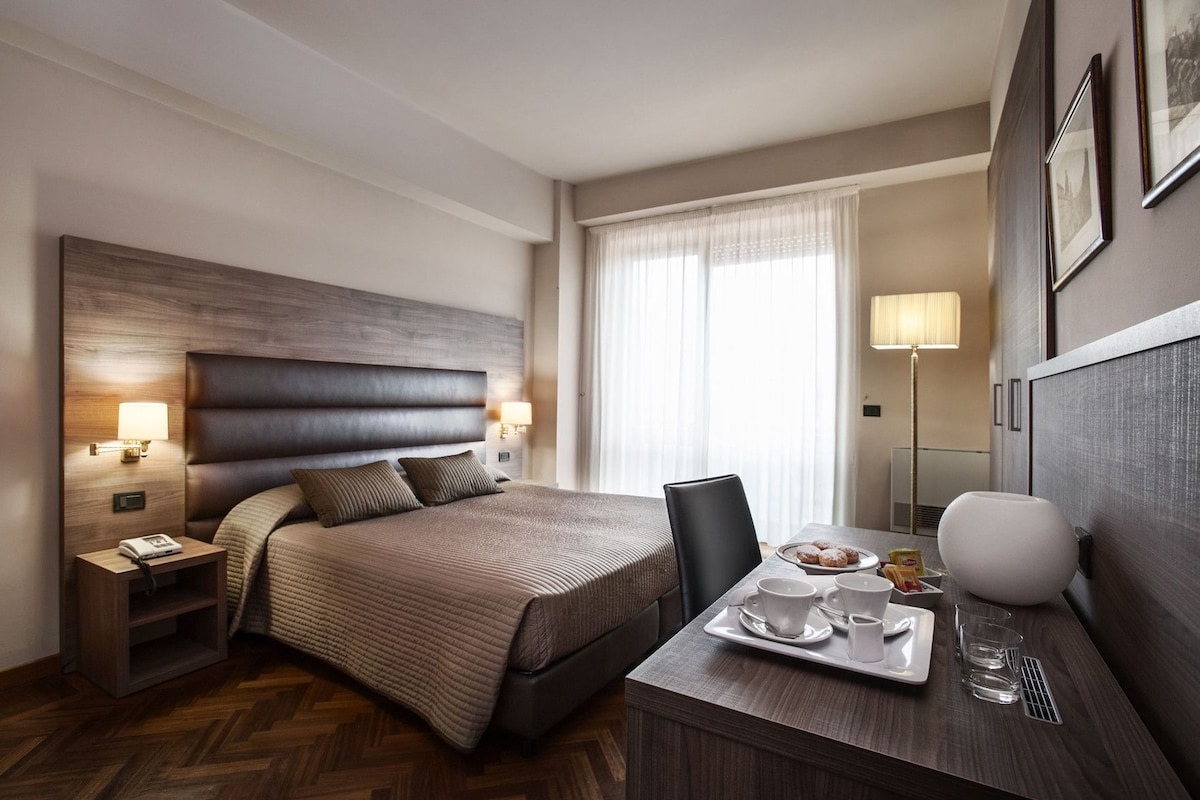 A hotel room with a bed, bedside tables, a desk with a tray of breakfast items, and a large window with curtains.