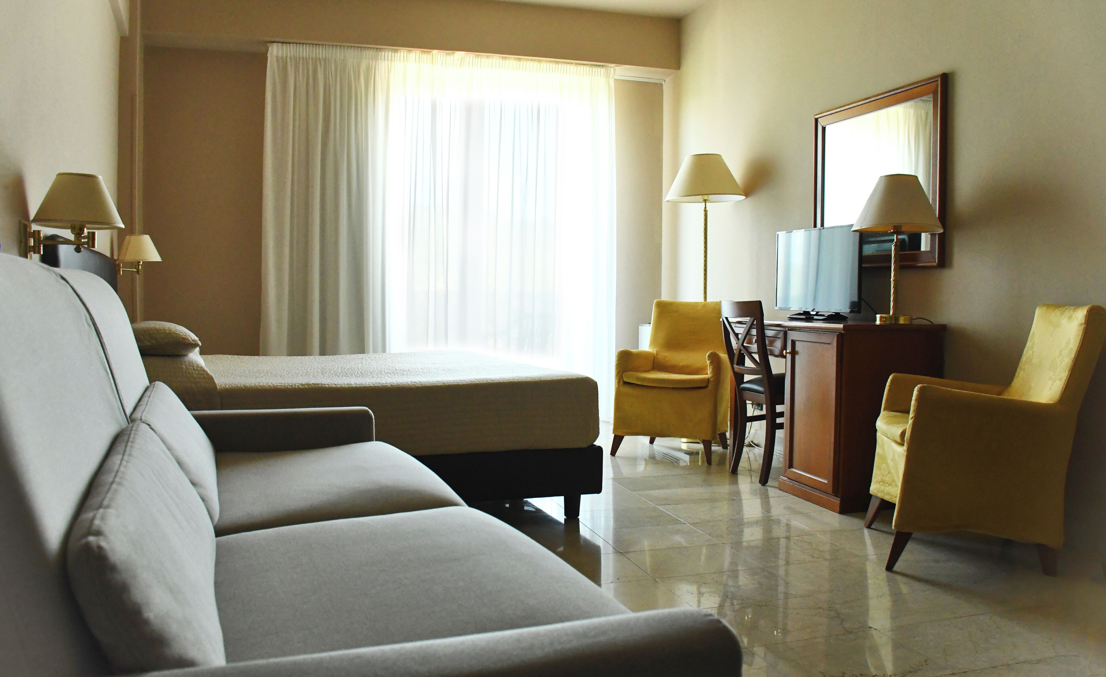 Habitación de hotel con cama, dos sillones, un escritorio con televisor y un ventanal con cortinas.