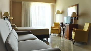 Habitación de hotel con cama, dos sillones, un escritorio con televisor y un ventanal con cortinas.