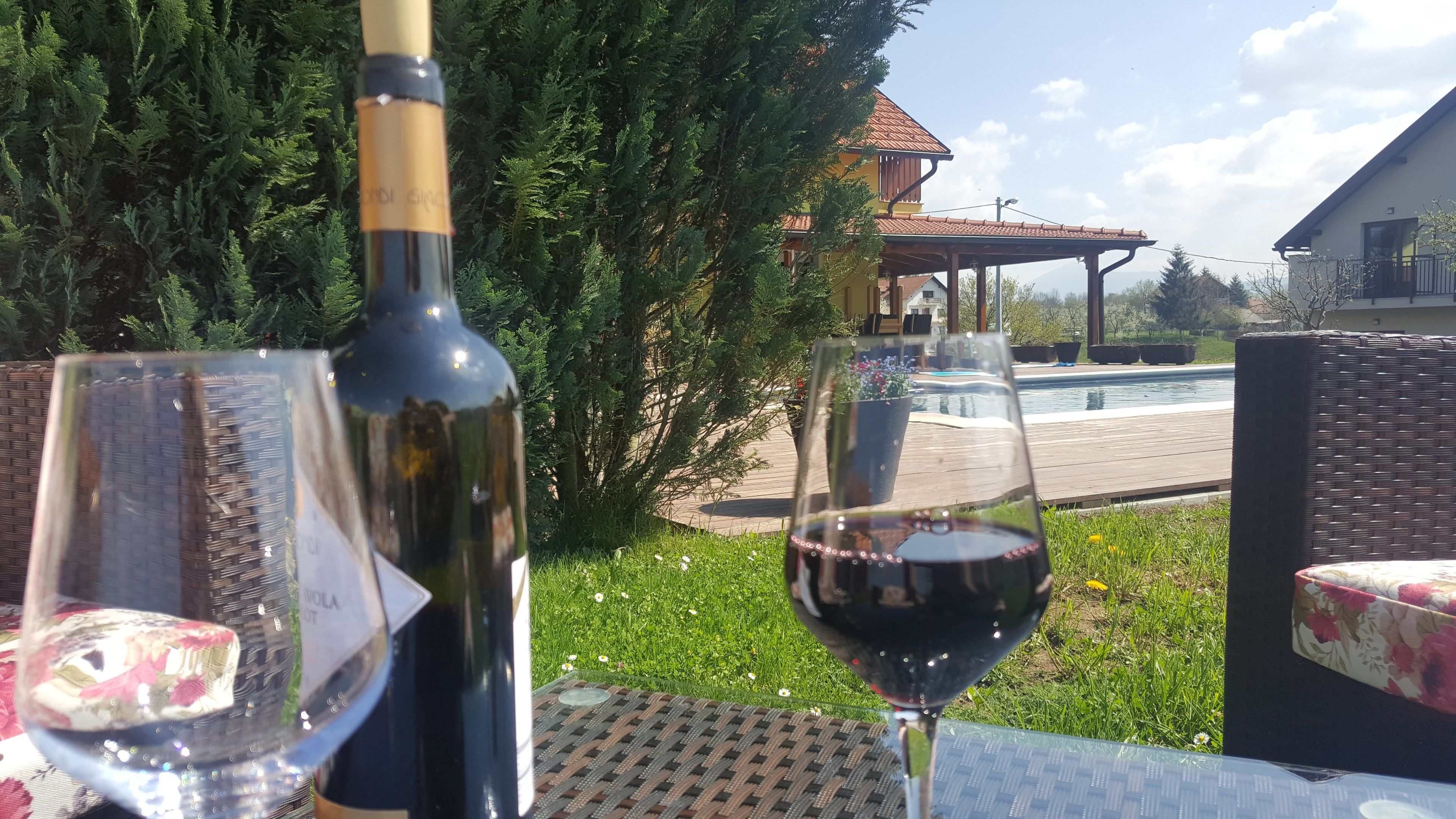 Una botella de vino y una copa de vino tinto sobre una mesa al aire libre, con vista a una piscina y una casa al fondo.