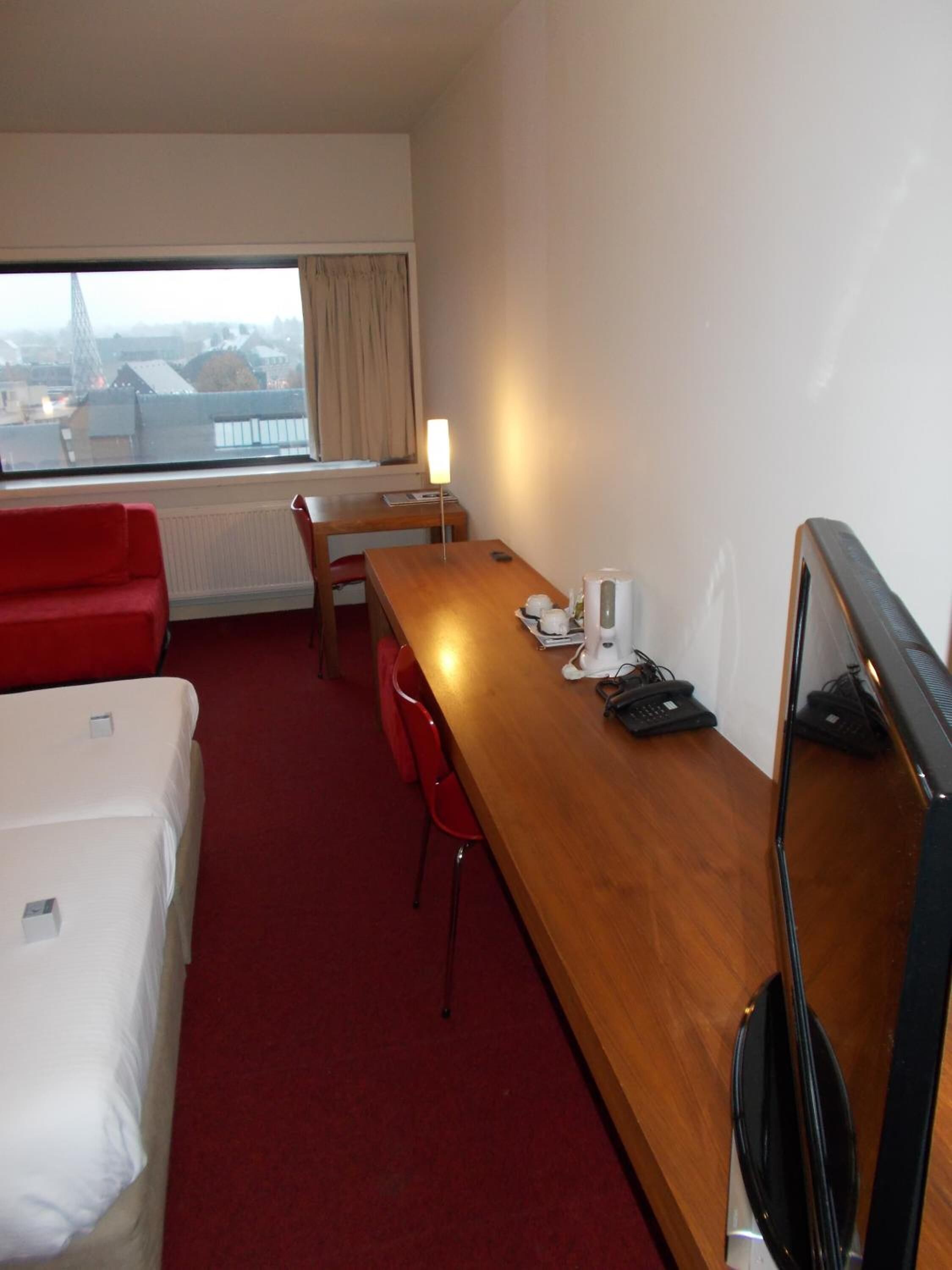A hotel room with a wooden desk, a red chair, a white bed, and a large window with a view of buildings.
