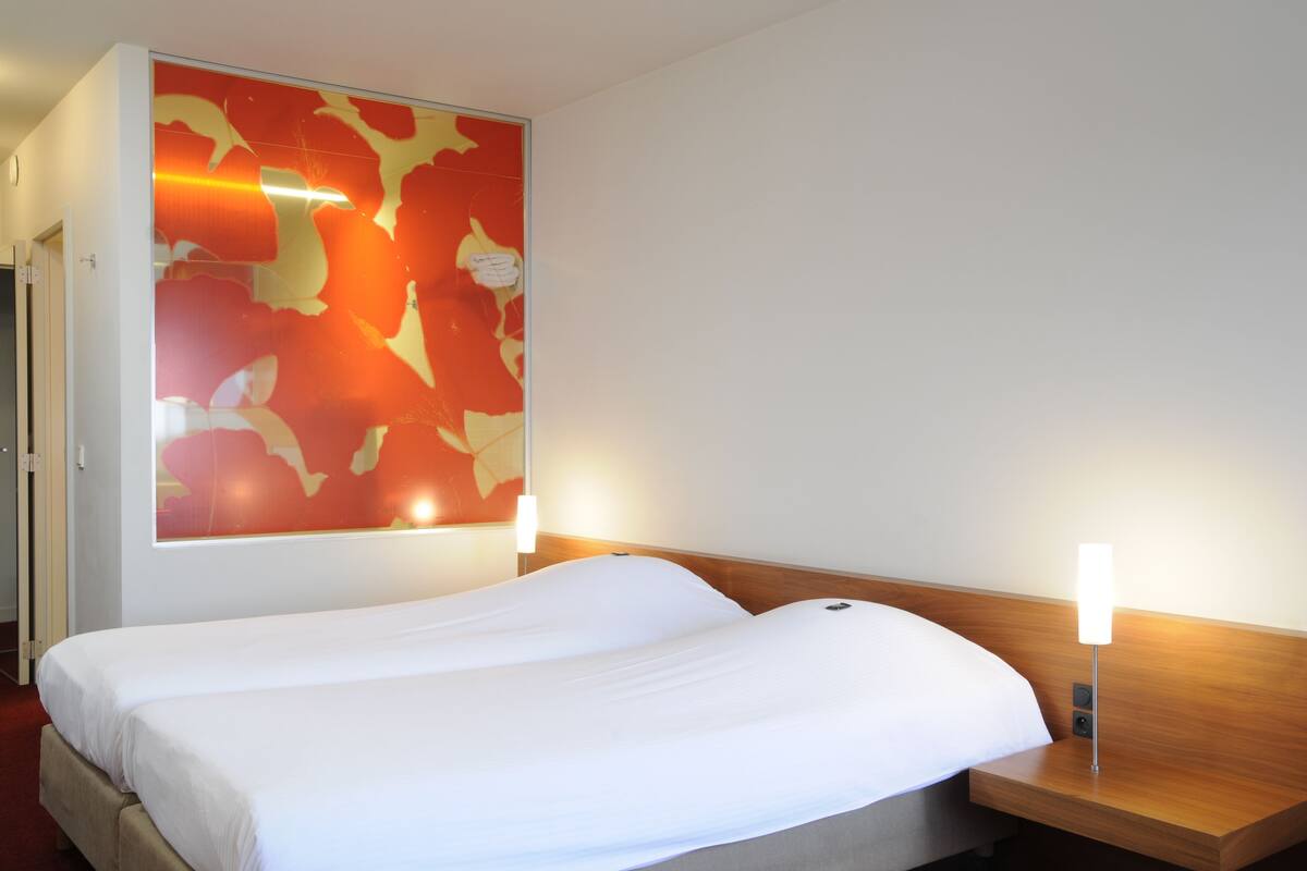 A hotel room with a bed, a wooden headboard, a wall with a red abstract design, and two bedside lamps.