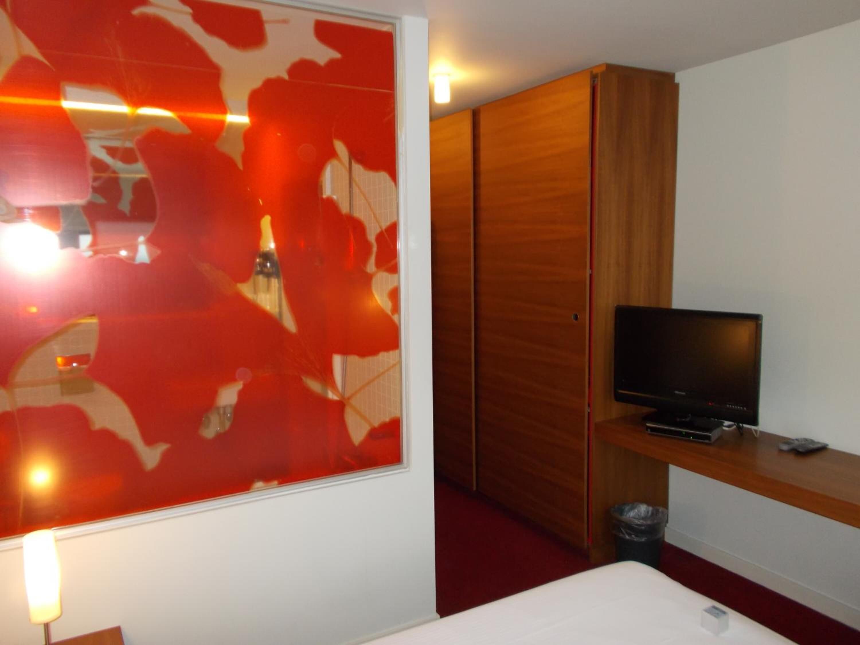 A hotel room with a large window, a television on a wooden shelf, and a red carpet.