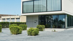 A modern hotel with a glass facade, a sign reading "Cordial Hotel," and a paved courtyard with neatly trimmed bushes.