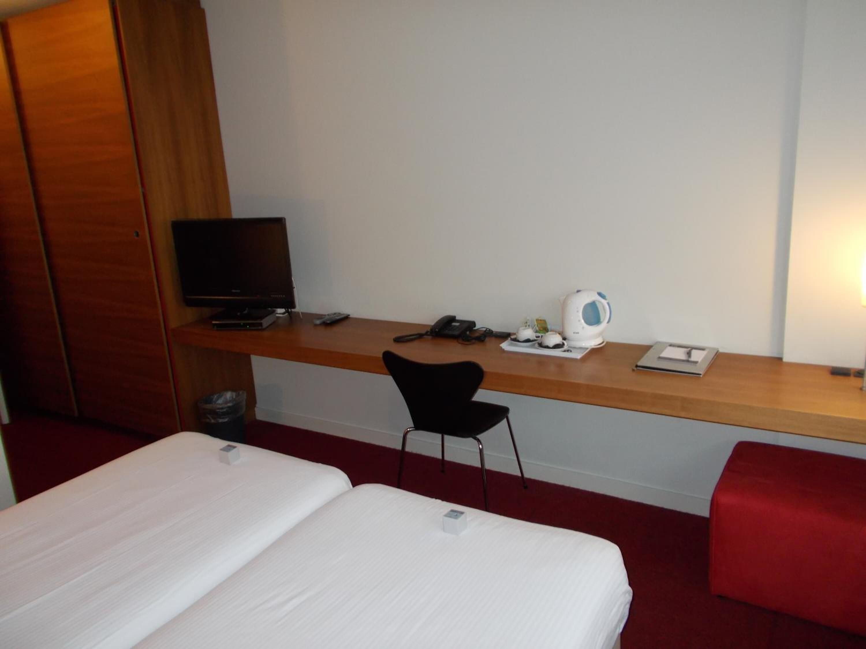 Habitación de hotel con una cama individual, un escritorio con computadora, una silla, un hervidor y una mesita de noche con lámpara.