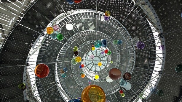 A spiral staircase with a glass enclosure, featuring colorful glass orbs suspended at various levels.