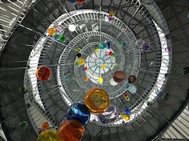 A spiral staircase with a glass enclosure, featuring colorful glass orbs suspended at various levels.
