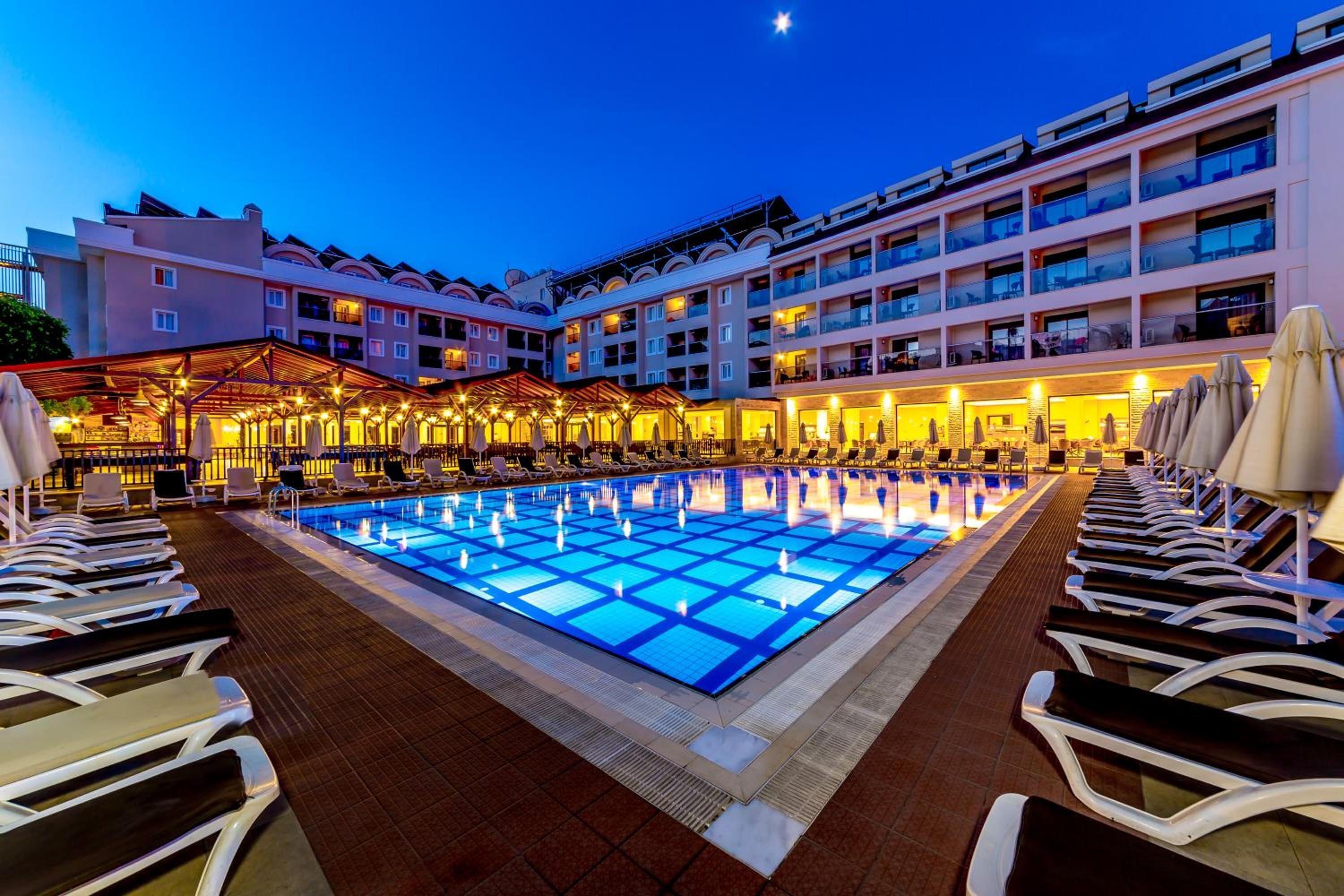 A hotel pool area with multiple lounge chairs and a large illuminated pool at dusk.