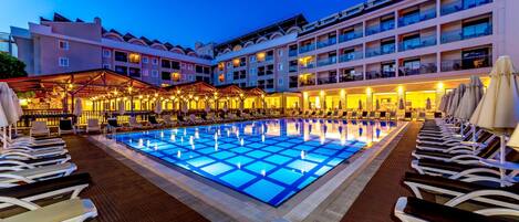 A hotel pool area with multiple lounge chairs and a large illuminated pool at dusk.