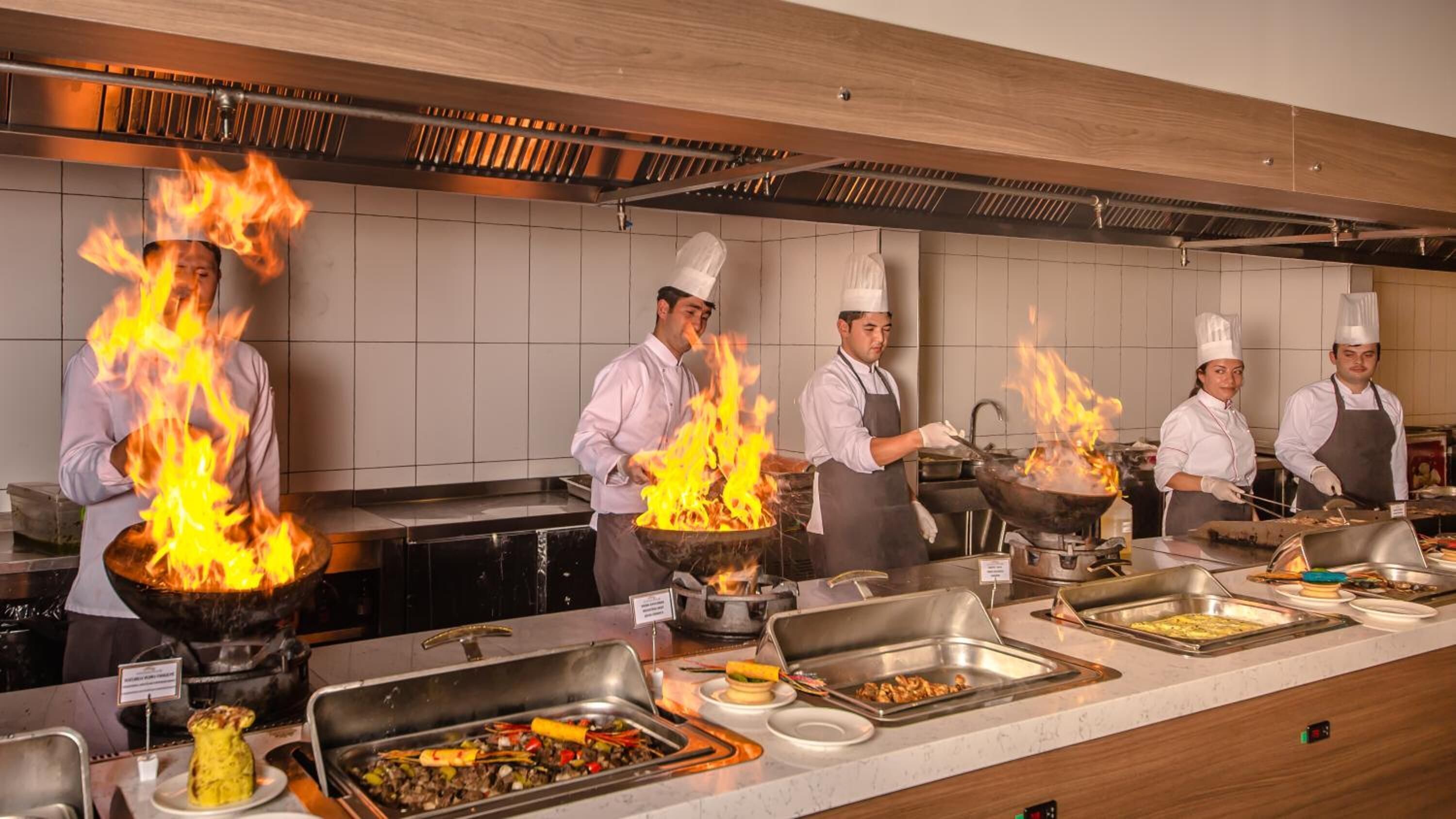 Una cocina profesional con chefs cocinando sobre fuego vivo, una barra de buffet con diversos platos, y un interior moderno y cuidado.