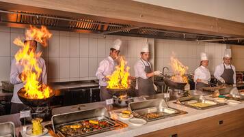 Una cocina profesional con chefs cocinando sobre fuego vivo, una barra de buffet con diversos platos, y un interior moderno y cuidado.