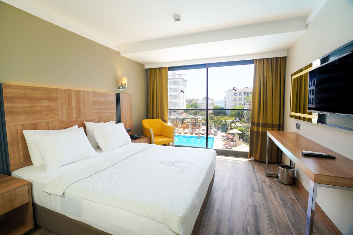 A hotel room with a large bed, a desk, a TV, and a view of a pool and buildings.