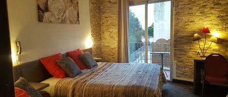 A bedroom with a large bed, a nightstand, a desk with a chair, a balcony with a railing, and a wall-mounted lamp.