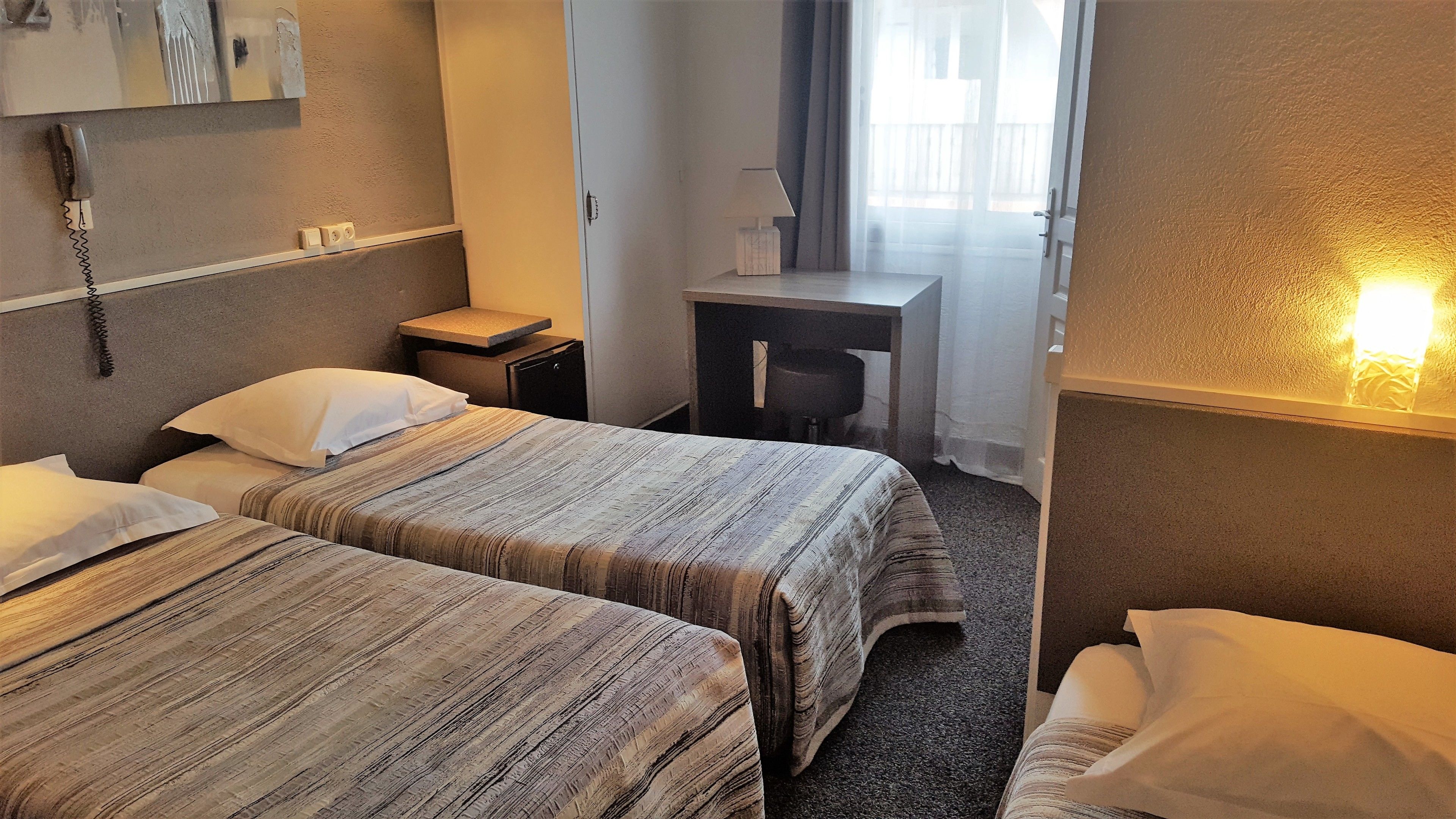 A hotel room with two beds, a desk, a chair, a lamp, and a phone on the wall.