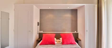 A hotel room with a bed, two red pillows, a headboard, a nightstand, a mirror, and a door.