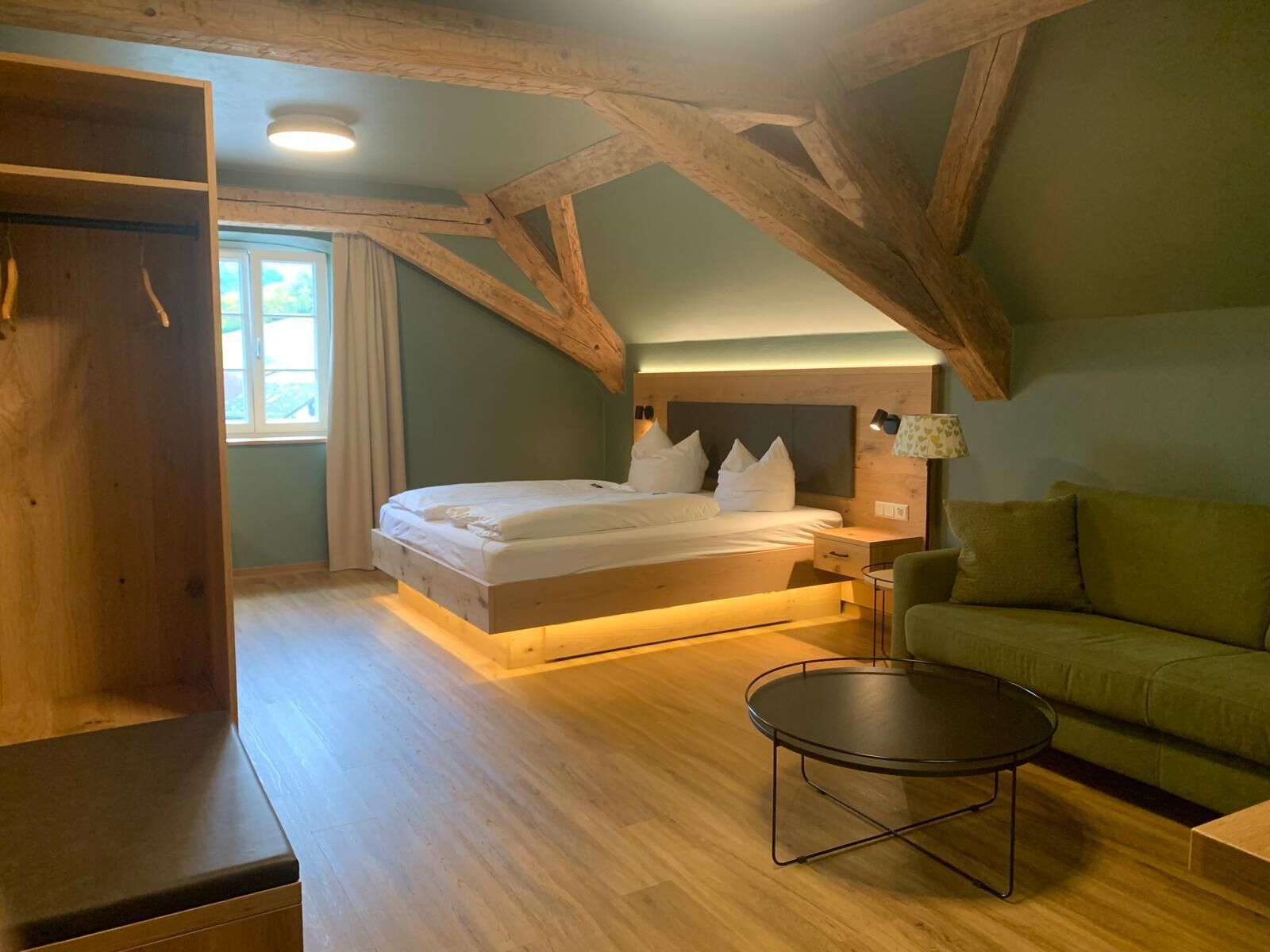 Un dormitorio con techo de madera, cama con ropa de cama blanca, mesita de noche con lámpara, sofá verde y una pequeña mesa redonda.