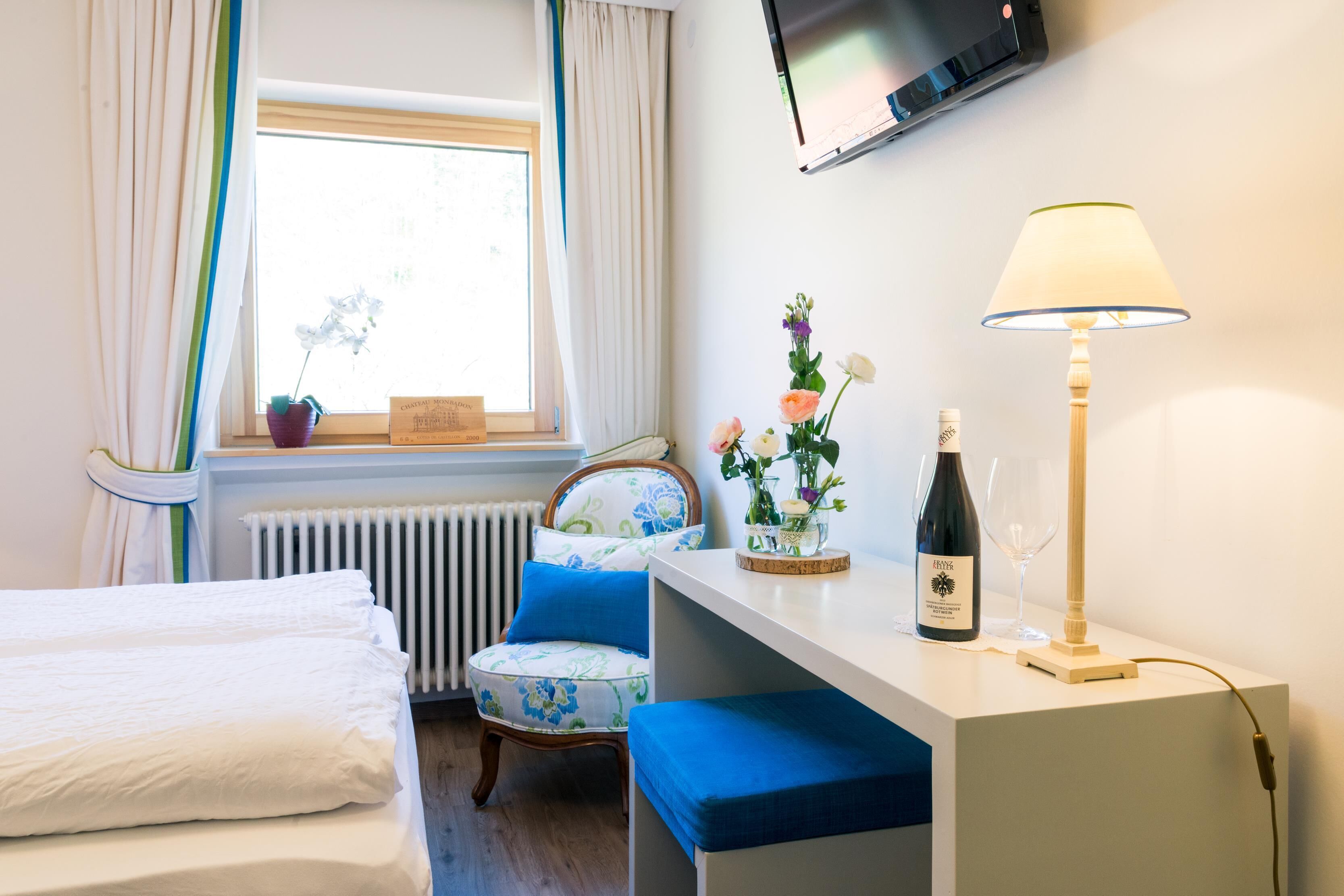 A hotel room with a bed, a chair, a desk with a lamp, a bottle of wine, and a TV on the wall.