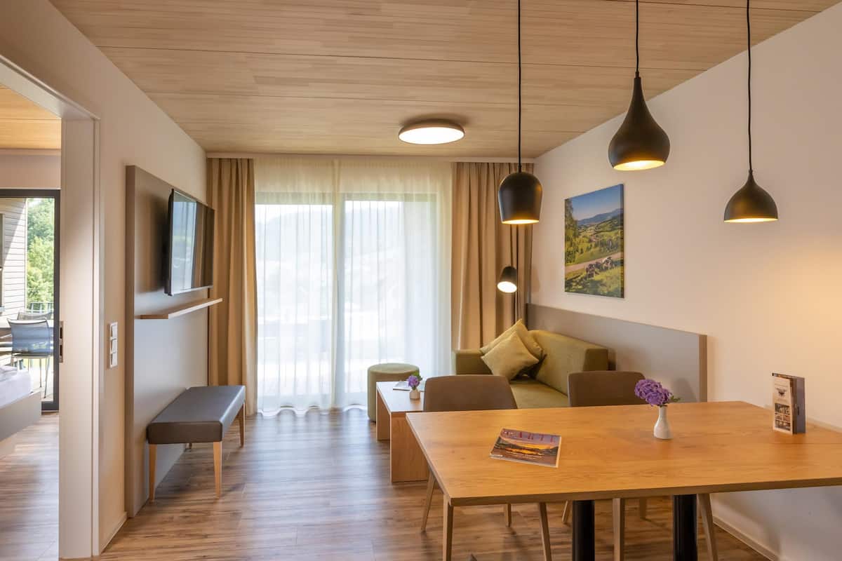 Ein modernes Hotelzimmer mit Holzboden, einem Esstisch, einer Couch, einer Bank und einem an der Wand montierten Fernseher.