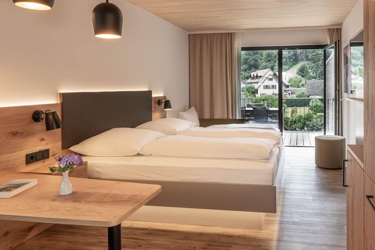 Ein modernes Hotelzimmer mit einem großen Bett, einem hölzernen Kopfteil, Nachttischlampen, einem kleinen Tisch mit Blumen und einem Balkon mit Blick auf die Landschaft.
