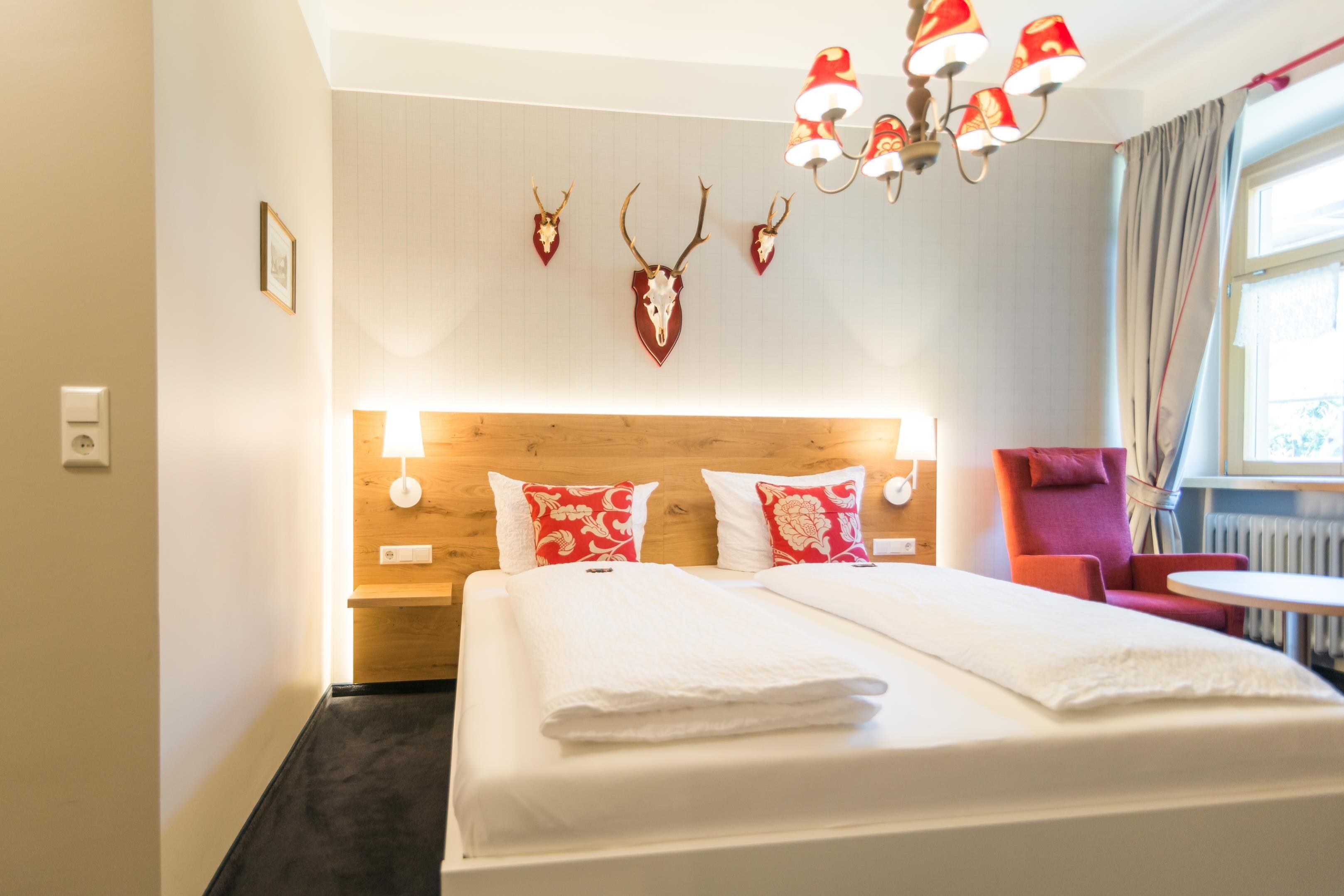 Une chambre d’hôtel avec une tête de lit en bois, deux coussins à motifs rouges et blancs, un couvre-lit blanc, une chaise rouge et un lustre.
