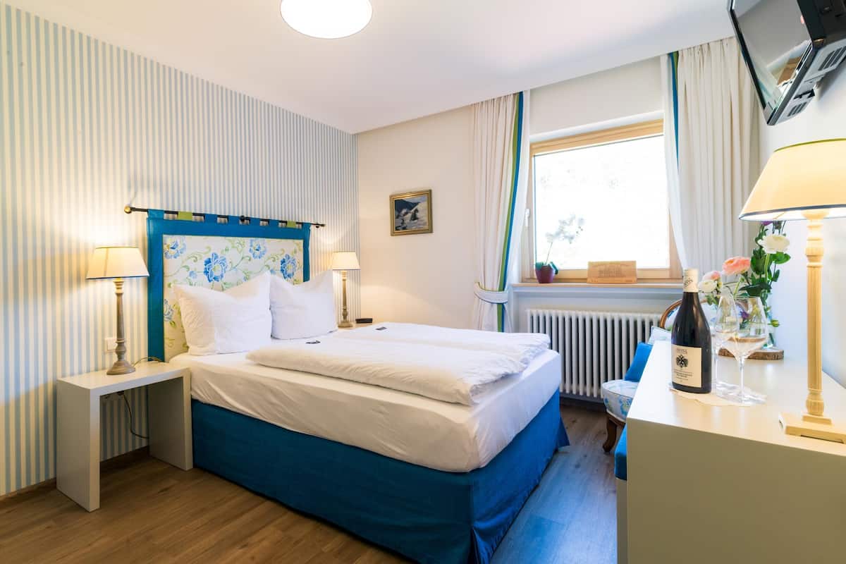 Ein ordentlich eingerichtetes Schlafzimmer mit Bett, Nachttischen, einem Fenster mit Vorhängen, einem Heizkörper und einem Schreibtisch mit einer Flasche und Gläsern.