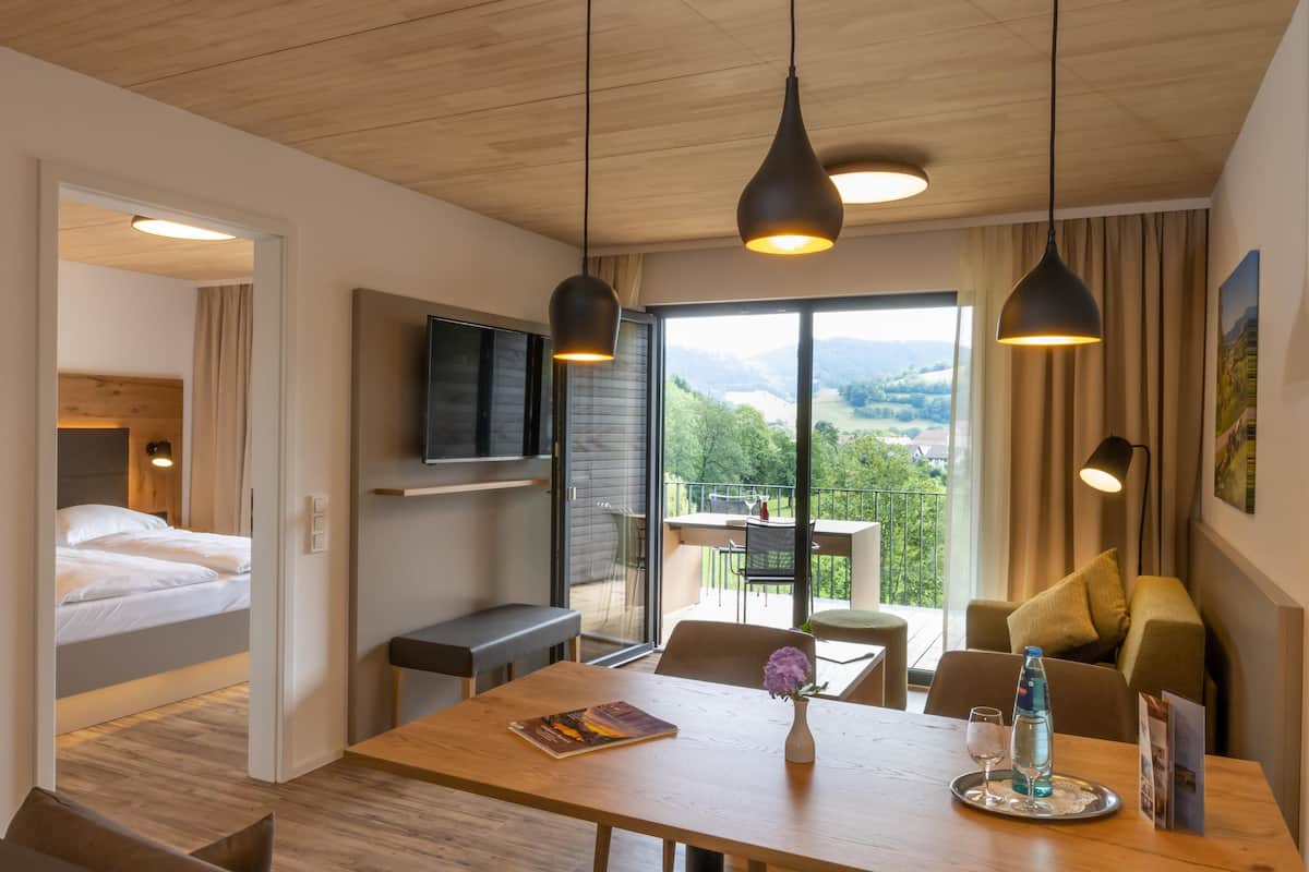 Ein modernes Hotelzimmer mit Holzboden, einem Bett, einem Essbereich mit Tisch und Stühlen sowie Blick ins Freie.