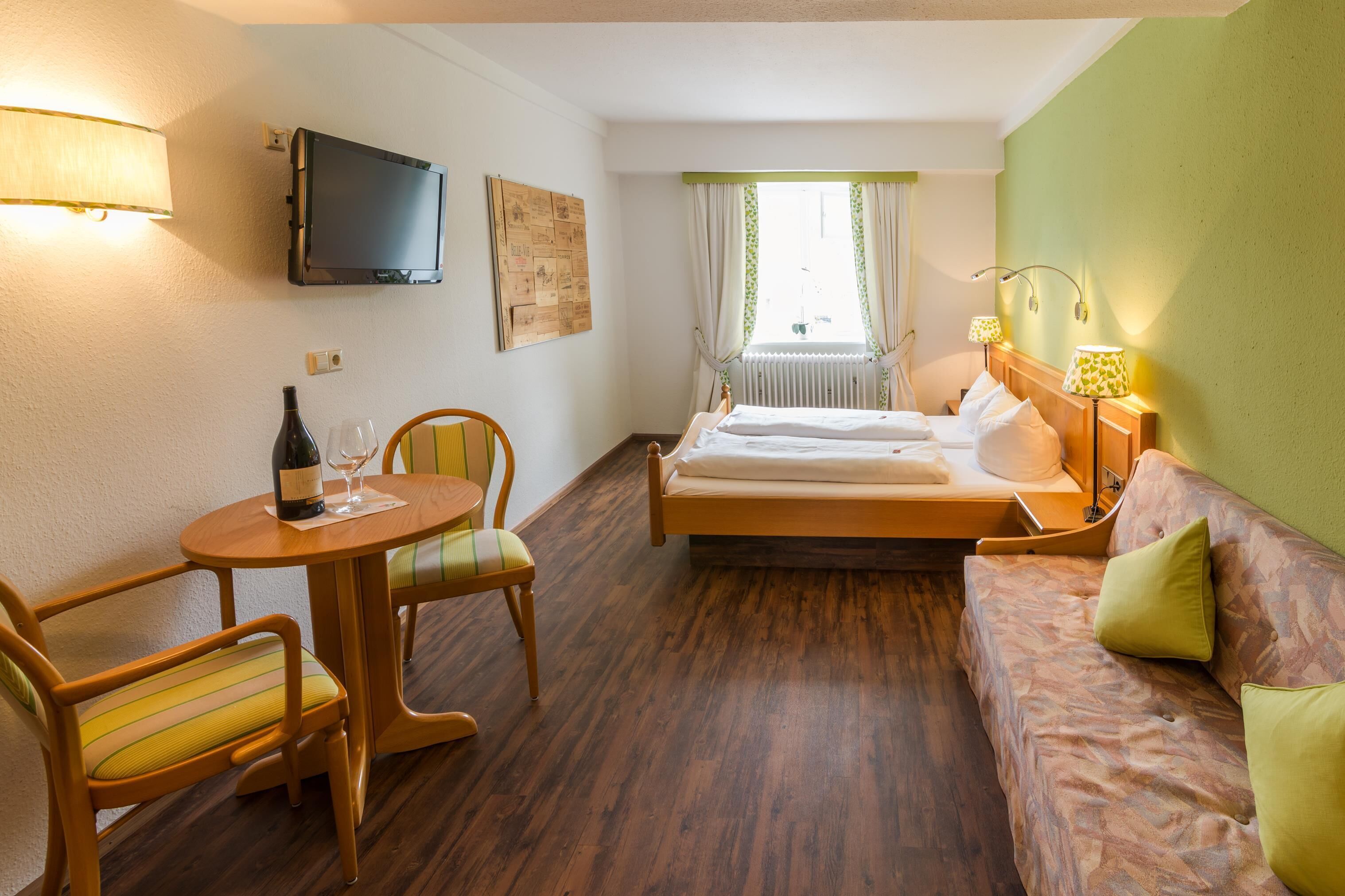 A hotel room with a bed, a sofa, a dining table with chairs, a TV mounted on the wall, and a window with curtains.