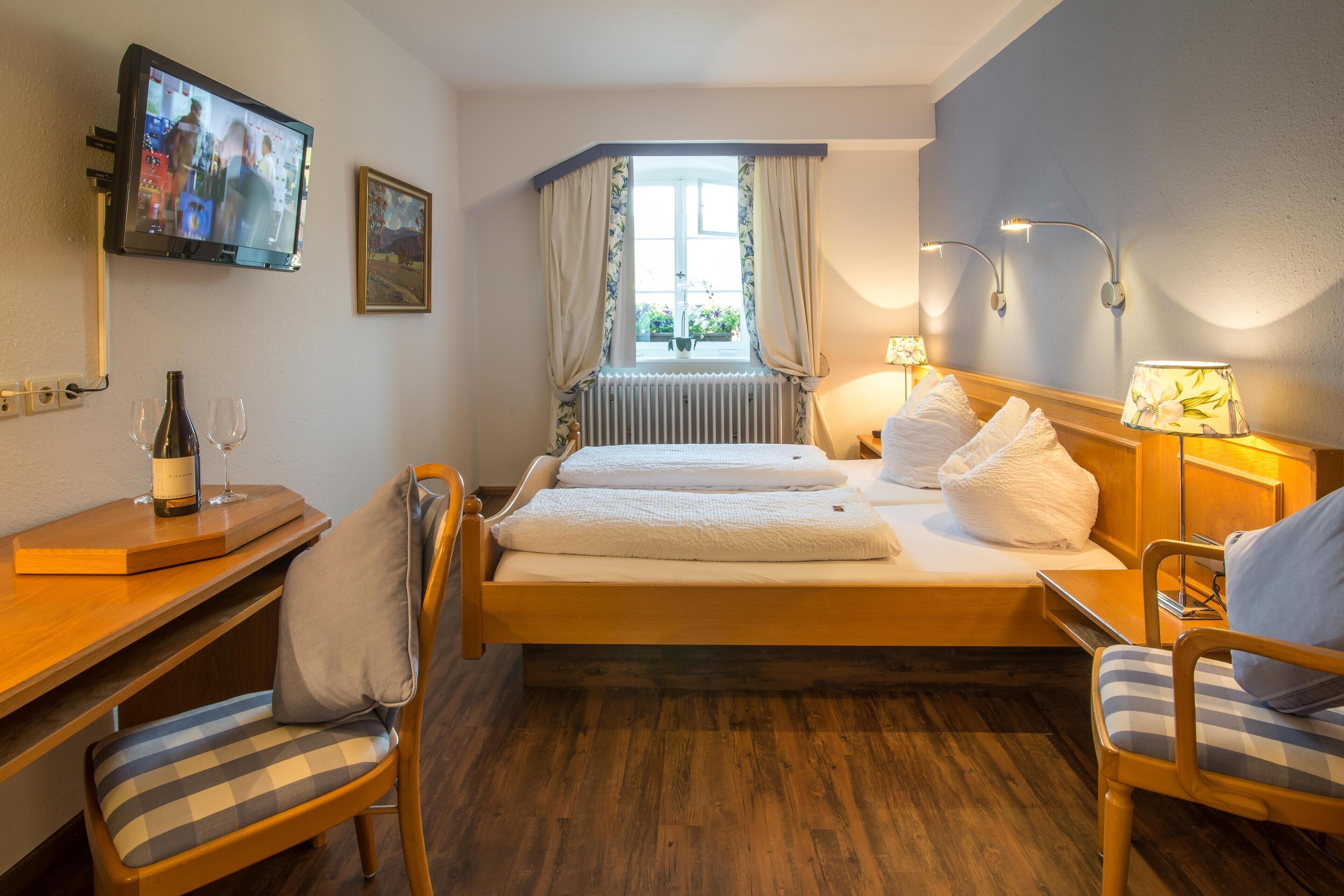 A hotel room with a bed, two chairs, a desk with a bottle and glasses, a TV, and a window with curtains.