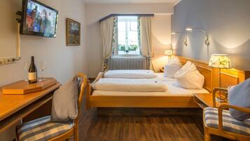 A hotel room with a bed, two chairs, a desk with a bottle and glasses, a TV, and a window with curtains.