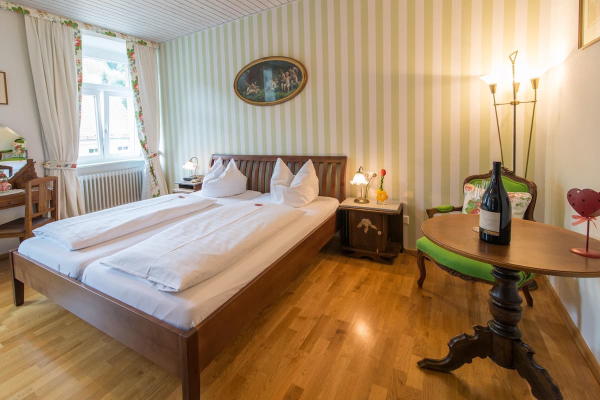 Ein Hotelzimmer mit einem großen Bett, Holzboden, einer grünen Polsterbank, einem kleinen Tisch mit einer Weinflasche und einem Wandbild.