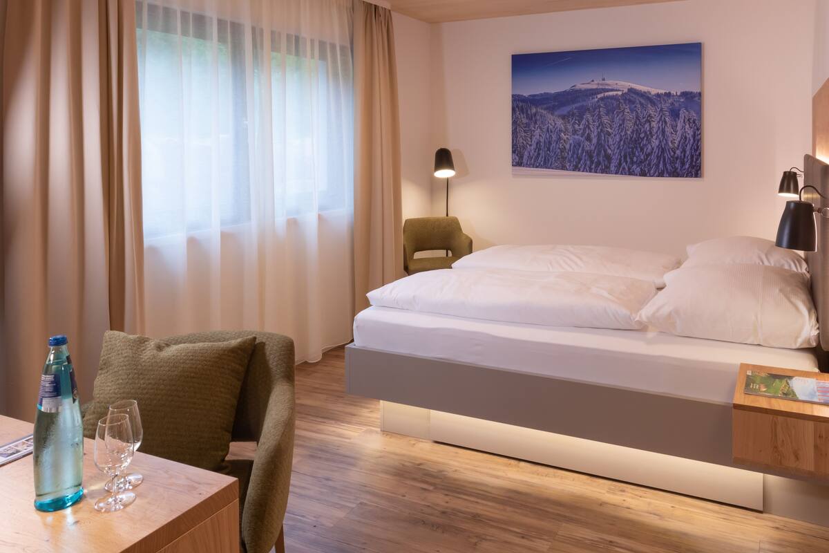 Ein modernes Hotelzimmer mit einem Bett, einem Sessel, einem Tisch mit Flasche und Gläsern, einer Lampe und einem Bild an der Wand.