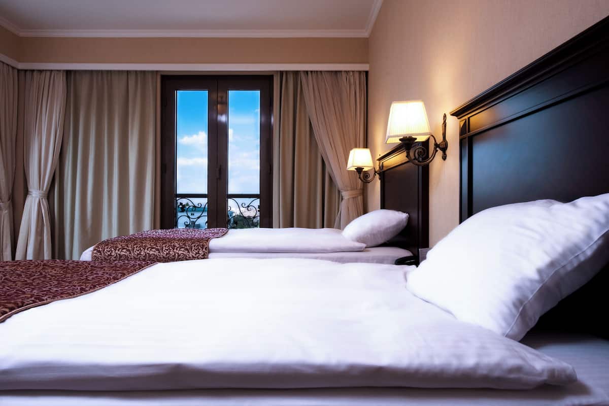 Ein Hotelzimmer mit zwei Betten, einem großen Fenster mit Vorhängen und einem Nachttisch mit Lampe.