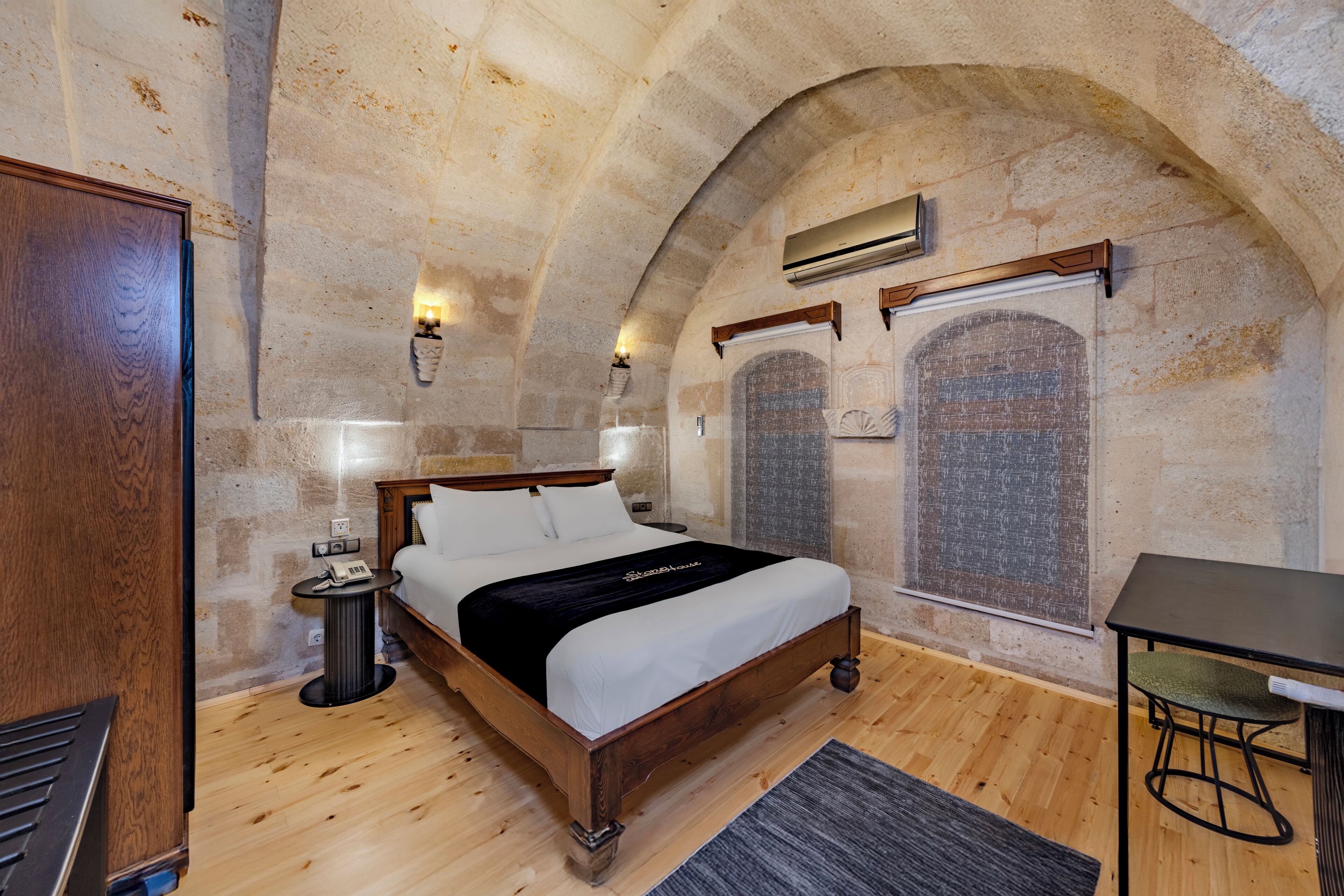 A bedroom with a wooden bed, a small bedside table, a wall-mounted air conditioner, and a stone wall.