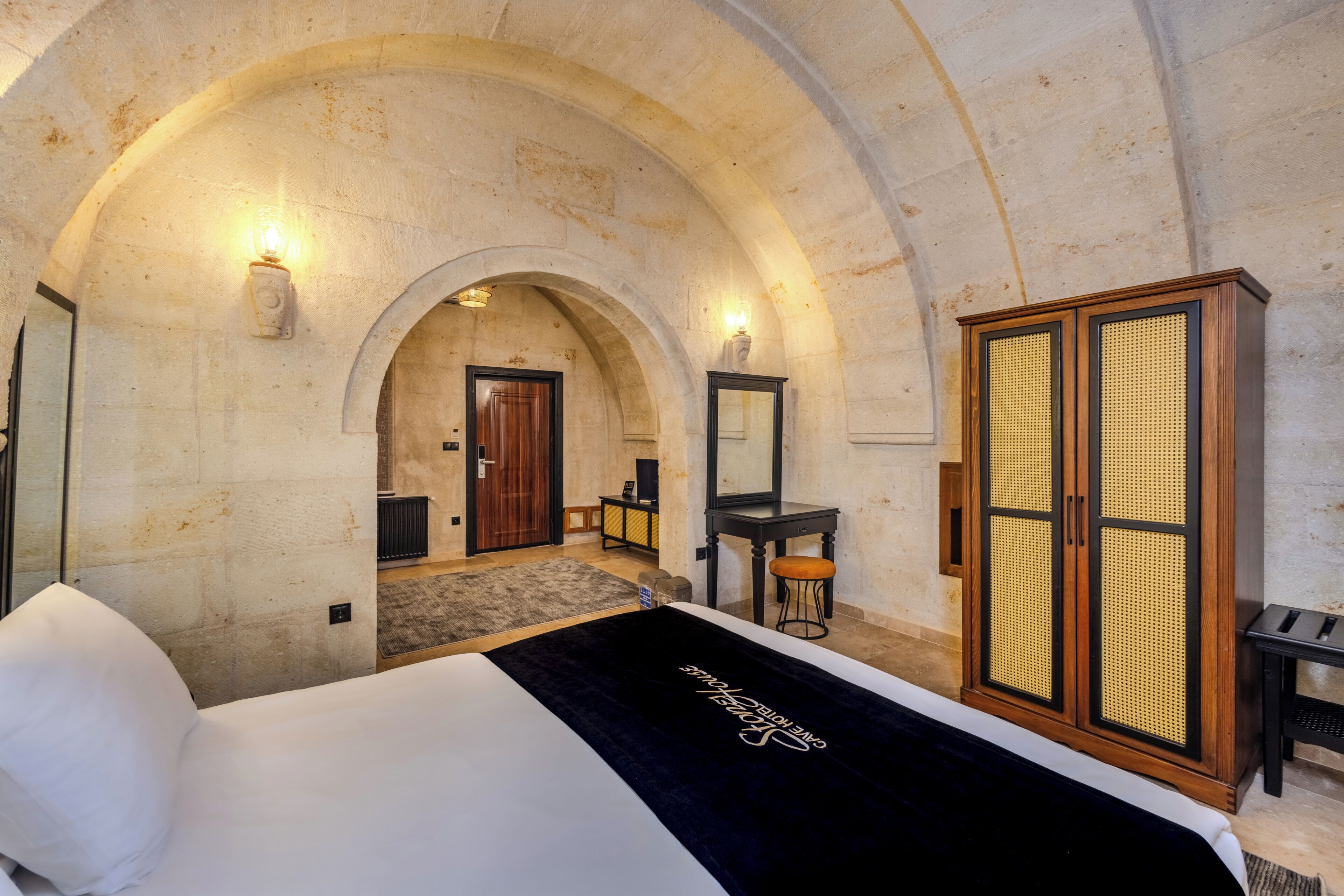 Une chambre d’hôtel comprenant un lit, une armoire, un miroir et une porte en arc donnant sur une autre pièce.