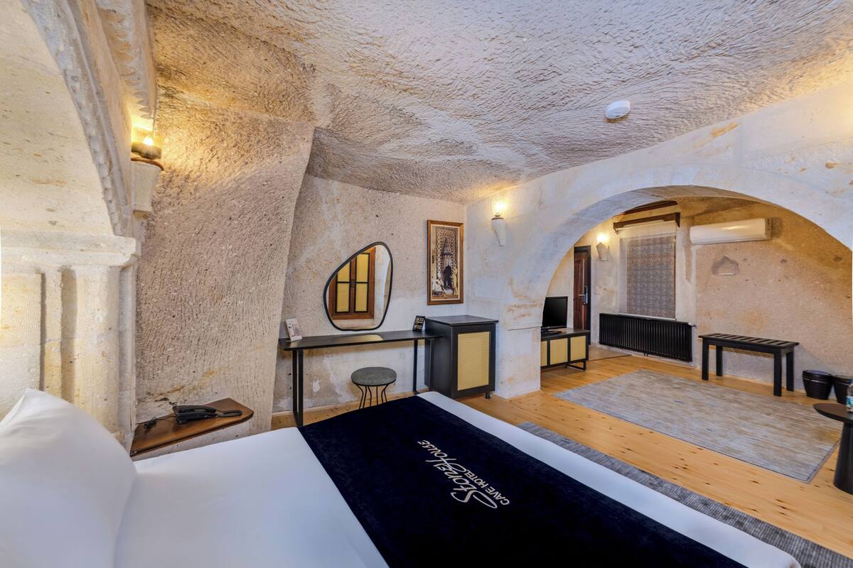 Ein Zimmer mit einem Bett, einem Schreibtisch, einem Stuhl und einem kleinen Hocker. An der Wand hängt ein Spiegel, und über dem Schreibtisch ein gerahmtes Bild. Das Zimmer hat eine rustikale Stein Decke und Wände.