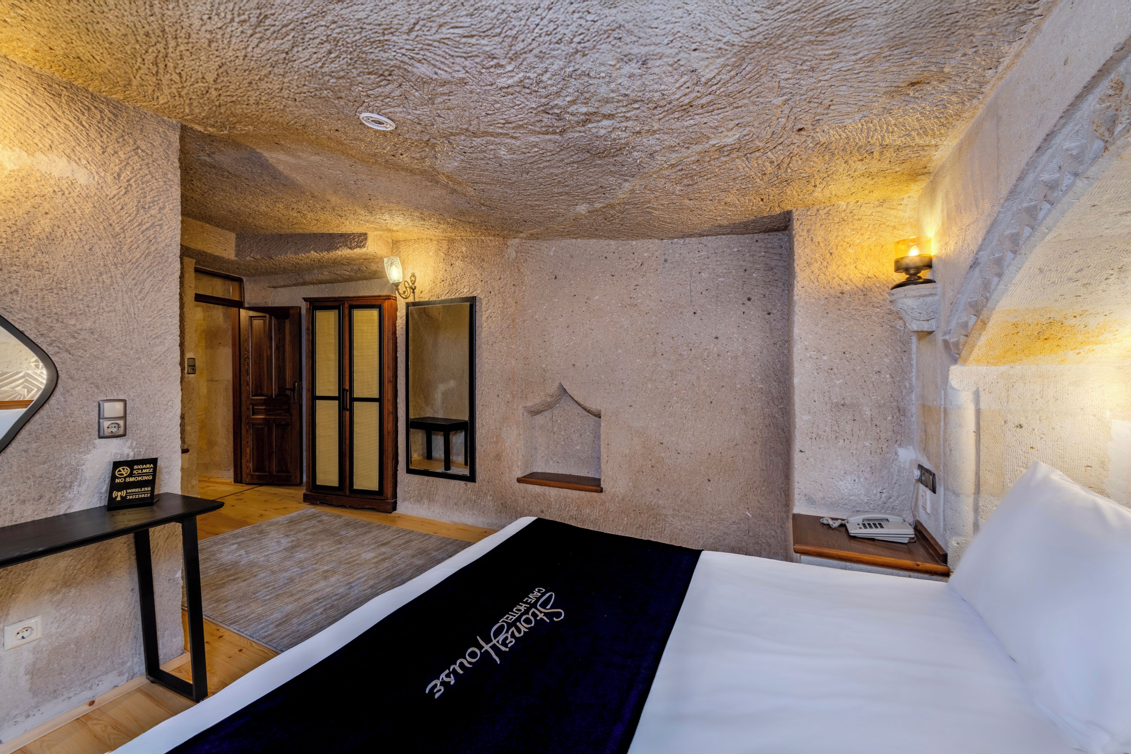 A bedroom with a bed, a bedside table, a mirror, and a door leading to another room.
