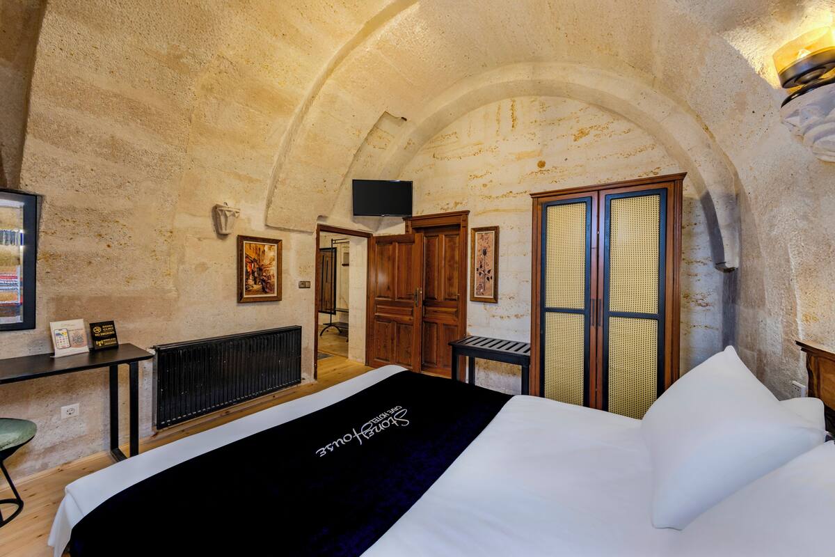 Ein Hotelzimmer mit einer Rundbogenzarge, einem Bett mit weißer Bettwäsche, einem Holztisch, einem Fernseher und Blick auf den Flur.