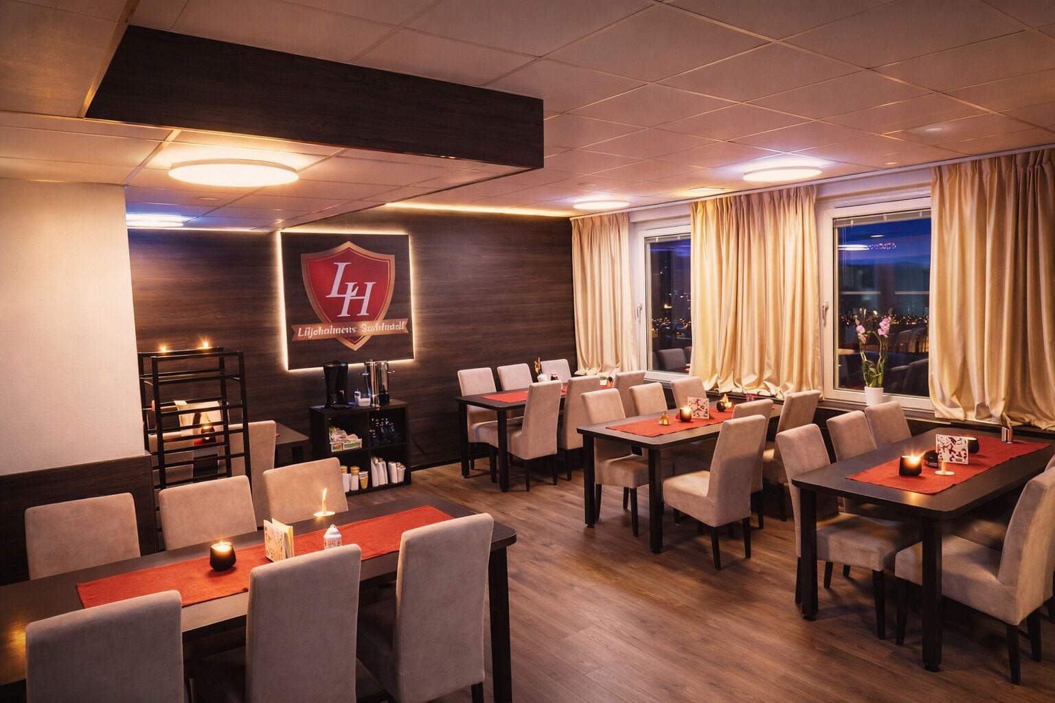 A hotel dining area with multiple tables and chairs, a large logo on the wall, and a view of the city at night through the windows.