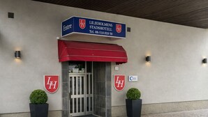 De ingang van Liljeholms Stadshotell met een rode luifel, twee potplanten en het logo van het hotel op de muur.
